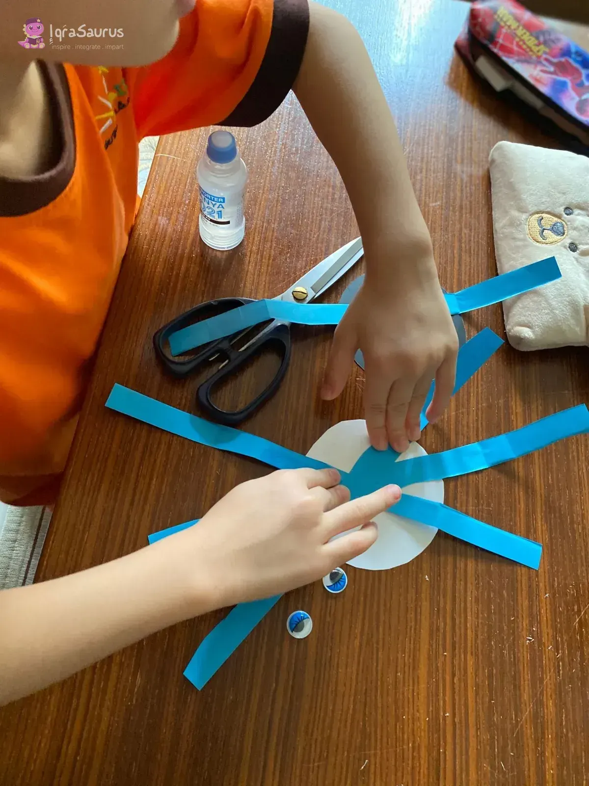 A child glues strips of blue paper onto a white paper circle to make spider legs, with googly eyes and scissors on the table